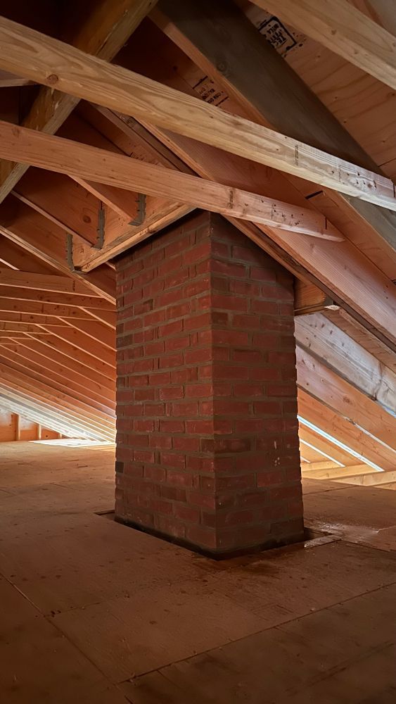 Brick chimney in attic, surrounded by wooden rafters. The chimney is rectangular and red. The attic has a wooden floor.