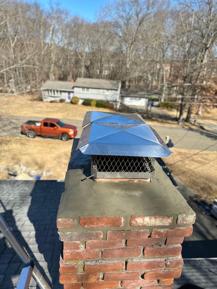 Chimney with a silver cap on a rooftop, orange truck in the background. Trees and houses in the distance on a sunny day.