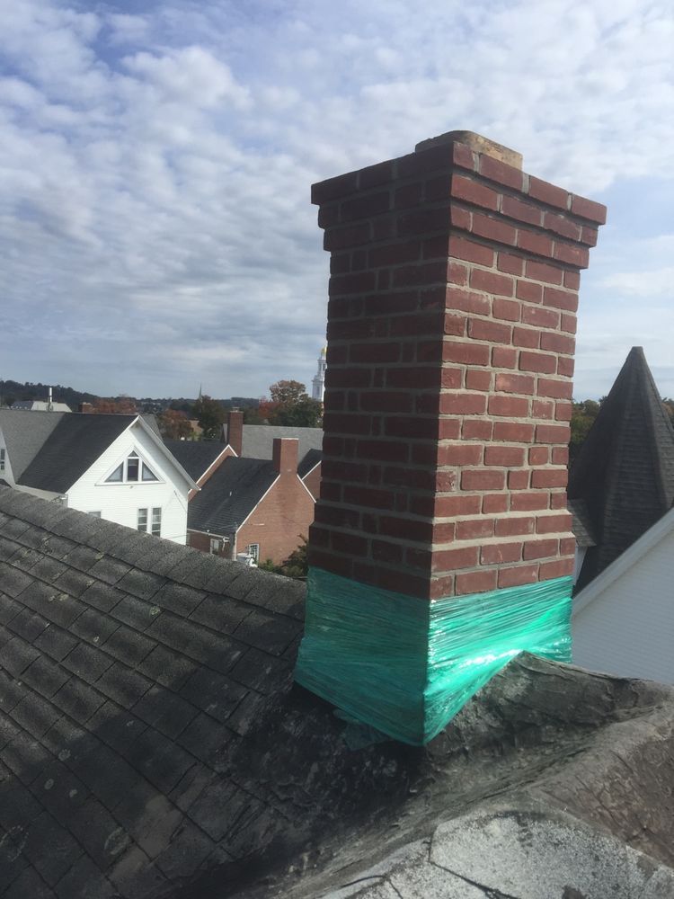 Brick chimney on a gray shingled roof, wrapped in green mesh. Houses and a cloudy sky are in the background.