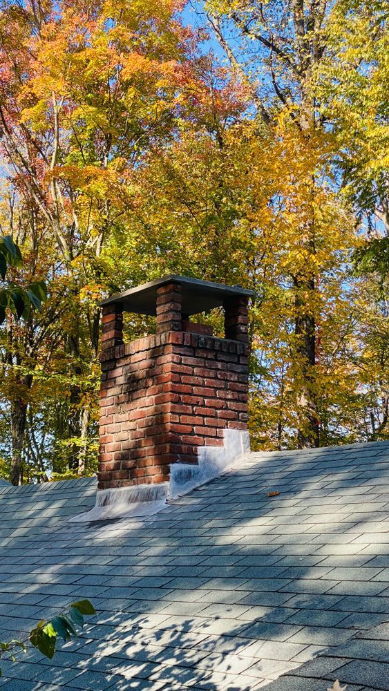 Brick chimney on a roof against a backdrop of trees with autumn foliage in yellow and orange.