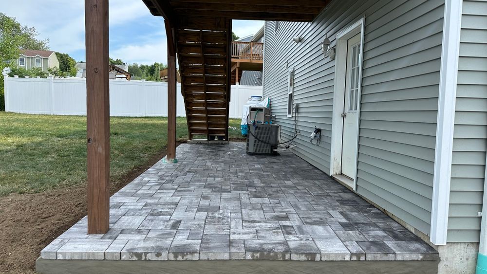 Patio with gray pavers, stairs leading up to a deck, next to a gray-sided house with a white door.