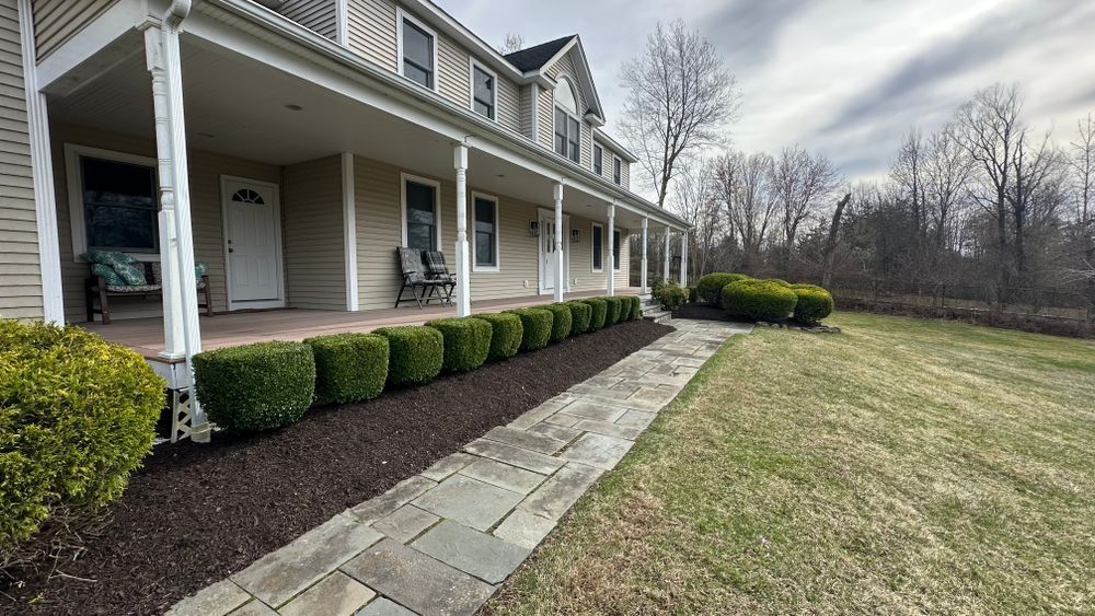A two-story beige house with a long porch. A stone walkway leads to the front door, lined with trimmed shrubs and brown mulch.