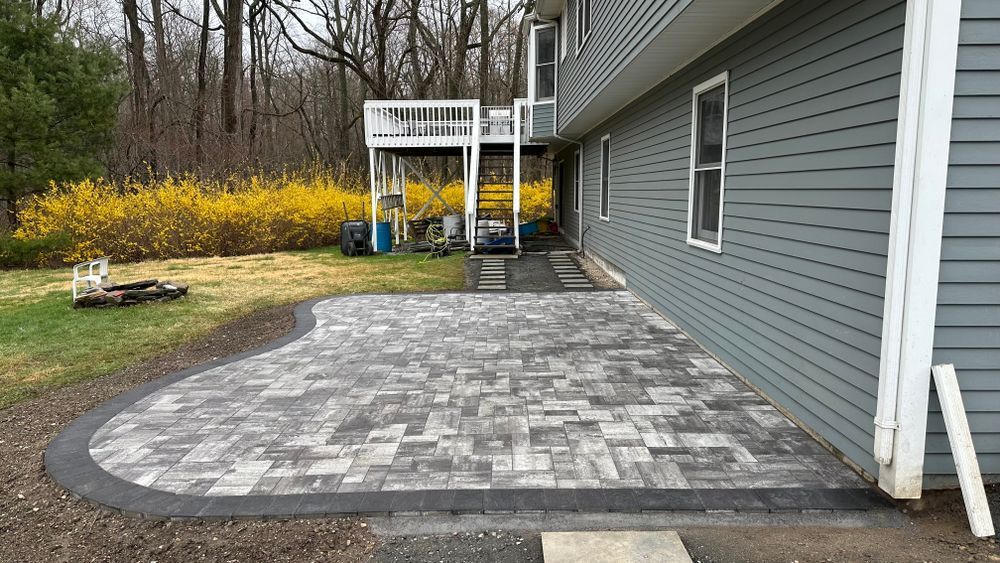 Gray paver patio next to a two-story gray house, with a raised deck in the background and yellow bushes on the left.