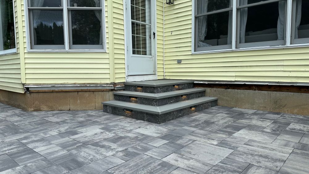 A set of three stone steps leading up to a white door on a yellow house. The steps are integrated into a gray brick patio.
