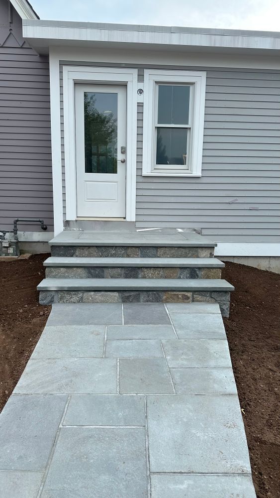 Gray stone walkway leading to a small building with a white door and window, accompanied by three steps of matching stone.