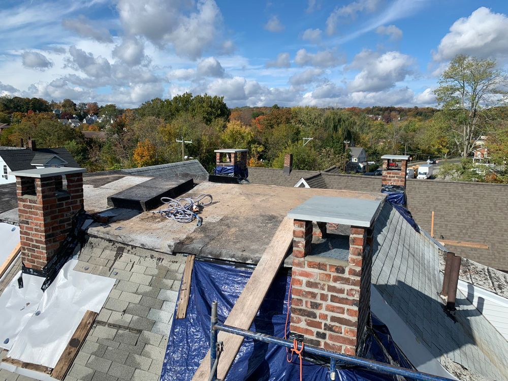 A rooftop with multiple brick chimneys. Construction in progress; tarps, exposed roofing material, and a blue sky.