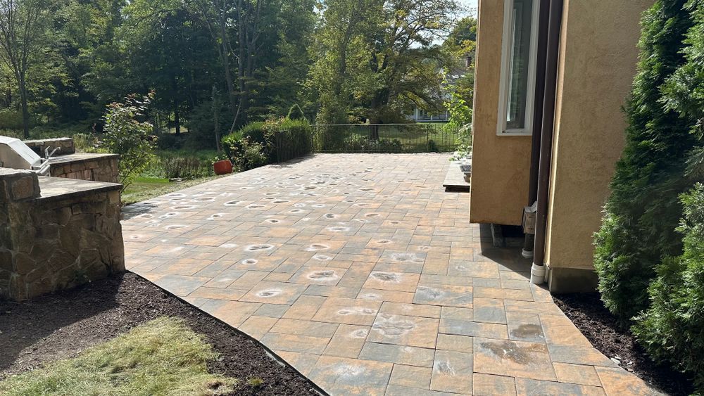 A paved patio with mixed-color stones next to a tan house and green landscaping. Trees are in the background.