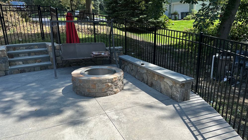 Patio with stone fire pit, benches, and metal fence. A swing and steps lead up to a higher level with a red umbrella.