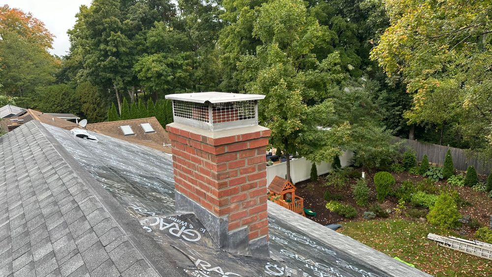 Brick chimney with a metal cap on a gray shingled roof against a background of green trees and a white fence.