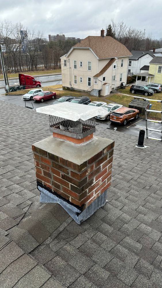 Brick chimney with a metal cap on a rooftop, surrounded by parked cars and buildings on a cloudy day.