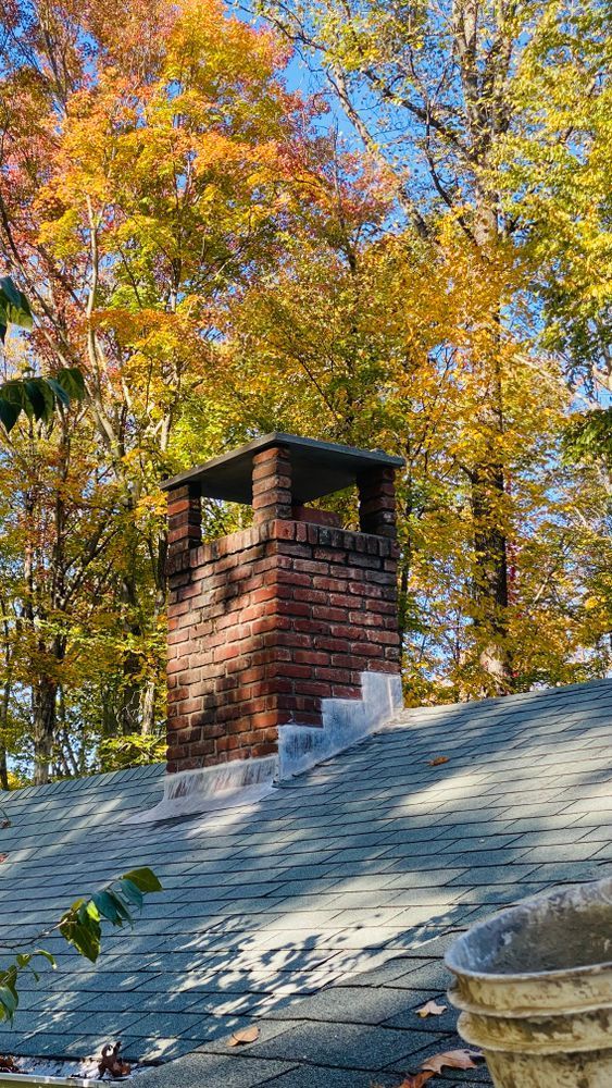 Brick chimney on a shingled roof with colorful autumn trees in the background under a blue sky.