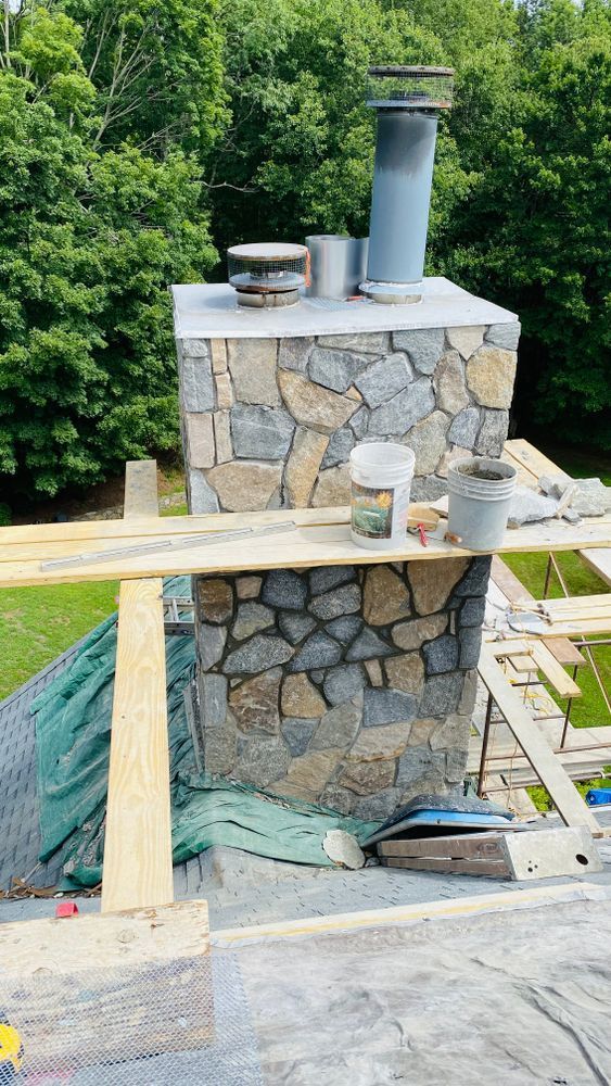 A stone chimney under construction on a roof, with wooden scaffolding. Green foliage and sky in the background.