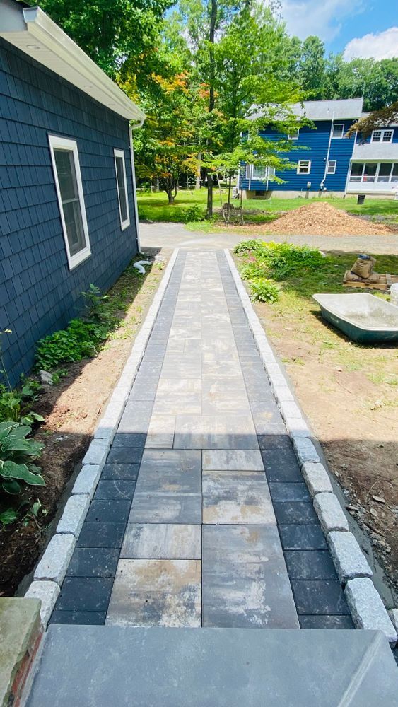 Brick walkway leading from blue house to a yard, lined with light gray pavers. Steps at the entrance.