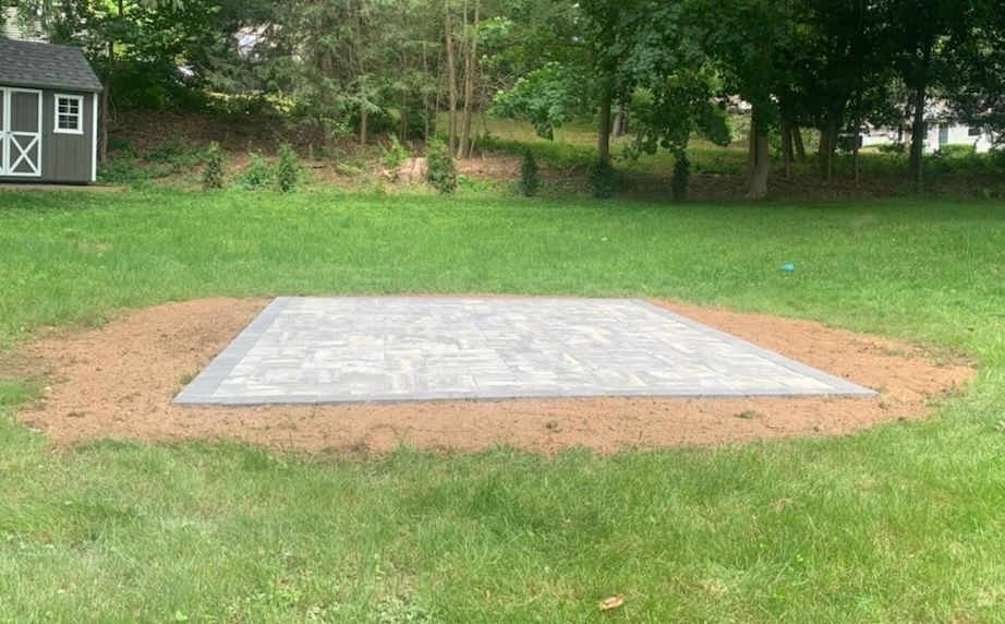 A completed stone patio, light gray, sits on a bed of brown soil in a grassy backyard. A shed is in the background.
