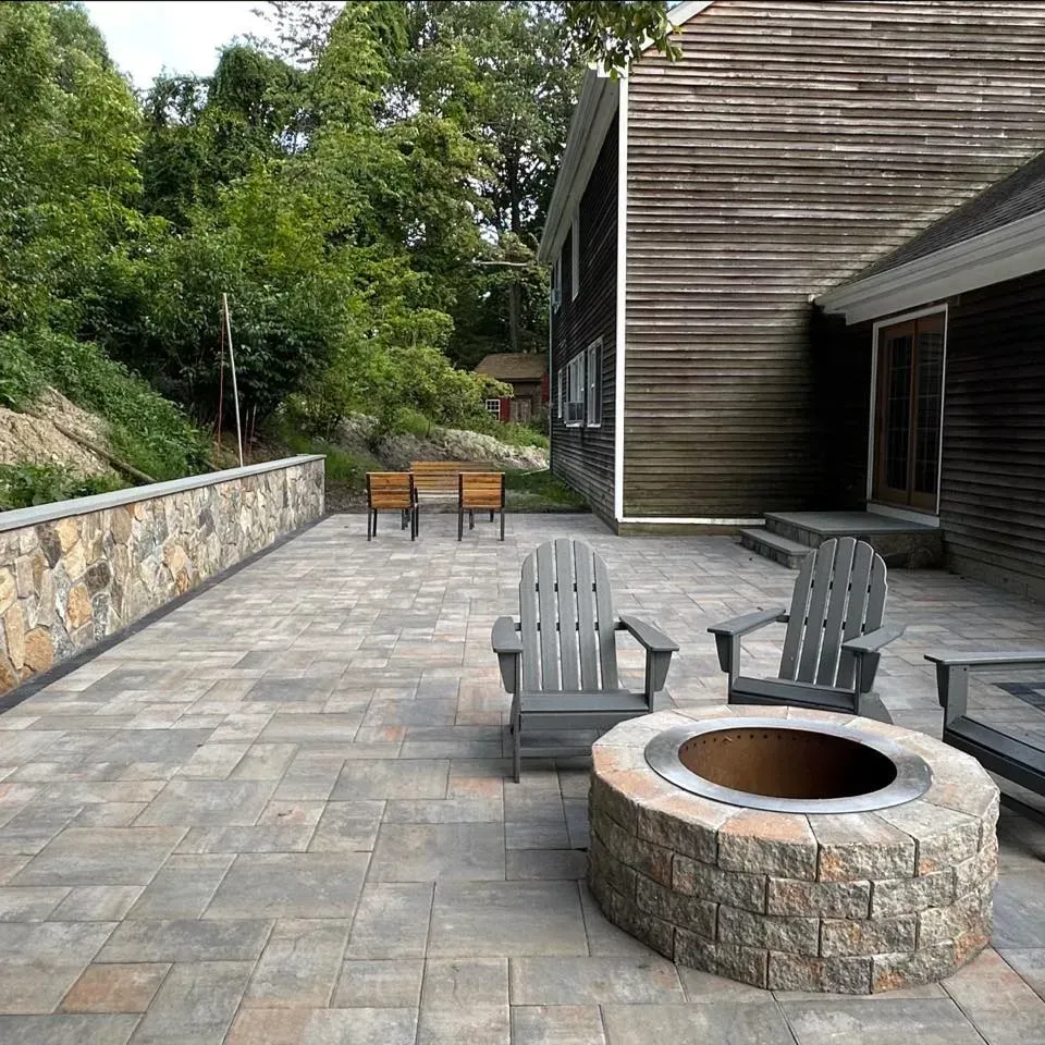 A stone patio with Adirondack chairs and a fire pit next to a wood-sided house and a stone retaining wall, surrounded by trees.