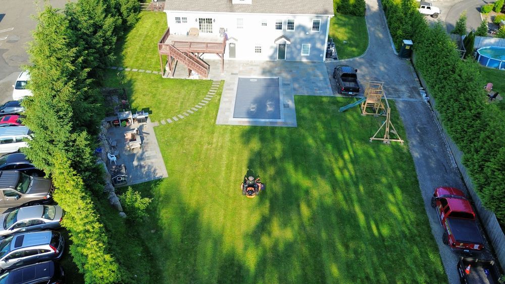Aerial view of a large house with a pool, lush green lawn, and a long driveway lined with cars and bushes.
