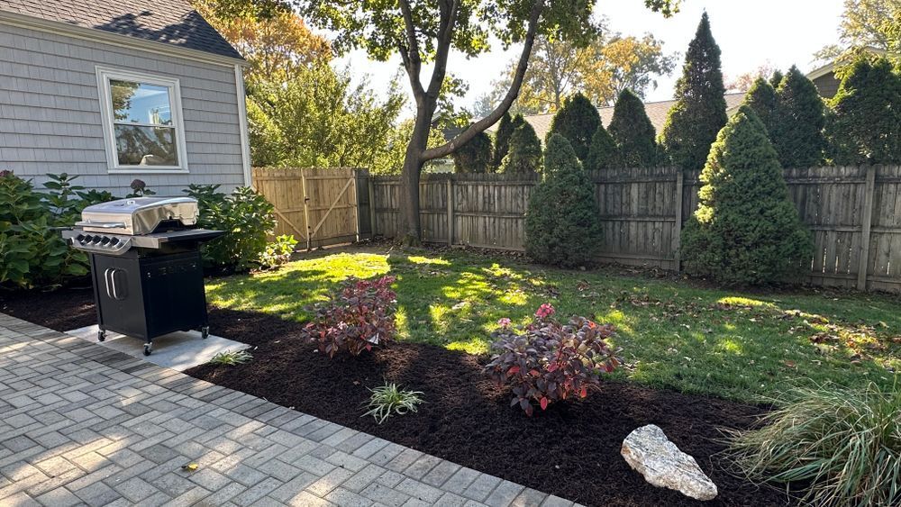 Backyard scene with a grill on a brick patio, mulched garden beds, and a wooden fence with evergreen trees.