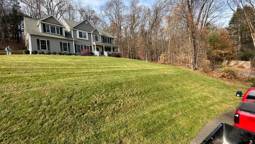 A two-story gray house with black shutters sits atop a grassy hill. A red pickup truck is in the foreground.