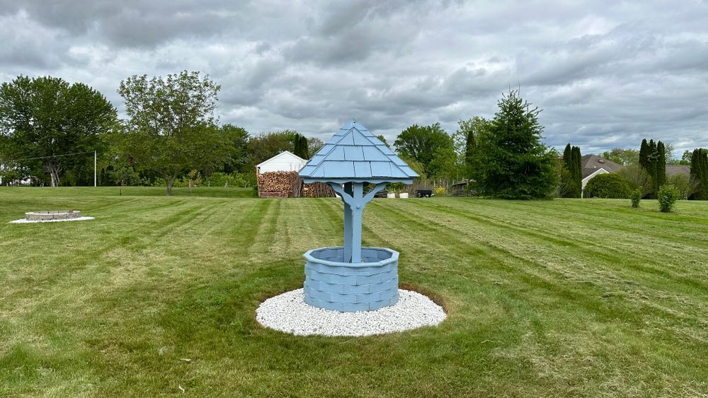 A blue wishing well with a white rock border sits on a grassy lawn under a cloudy sky.