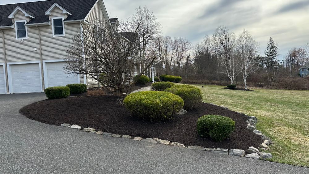 Curved driveway bed with dark mulch, green shrubs, and trees in front of a two-story house with white garage doors.