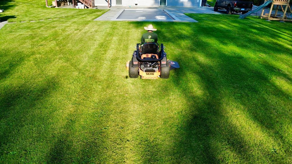 A person on a riding lawn mower cutting a large green lawn on a sunny day. The mower is yellow and black.