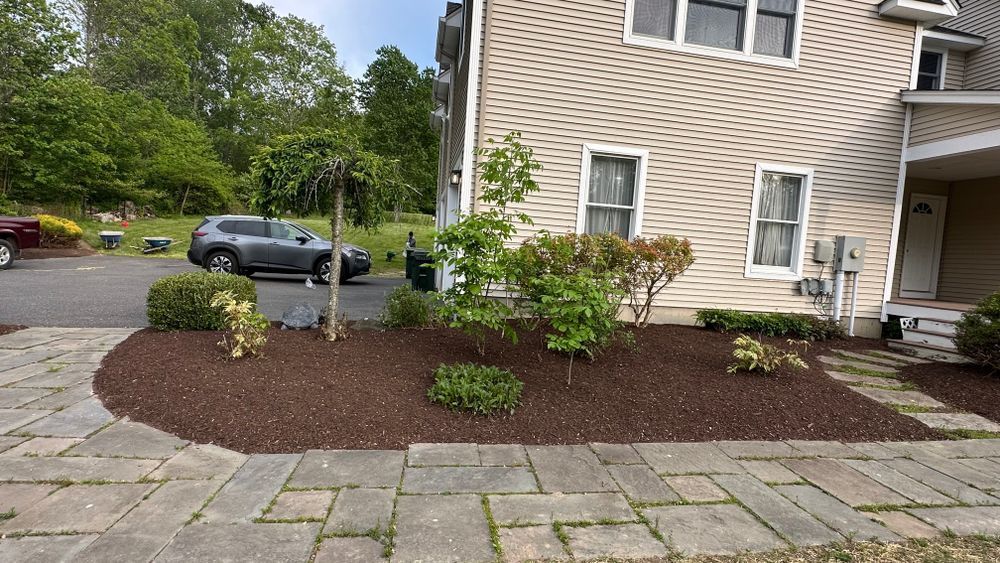 A tan house with a landscaped front yard featuring mulch beds, bushes, and a paved walkway. A car is parked in the driveway.
