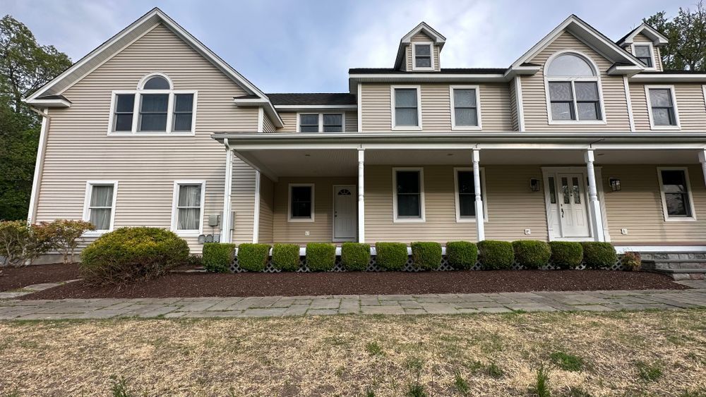 Beige two-story house with a porch, windows, and a dark roof. A line of green bushes borders the porch's base.