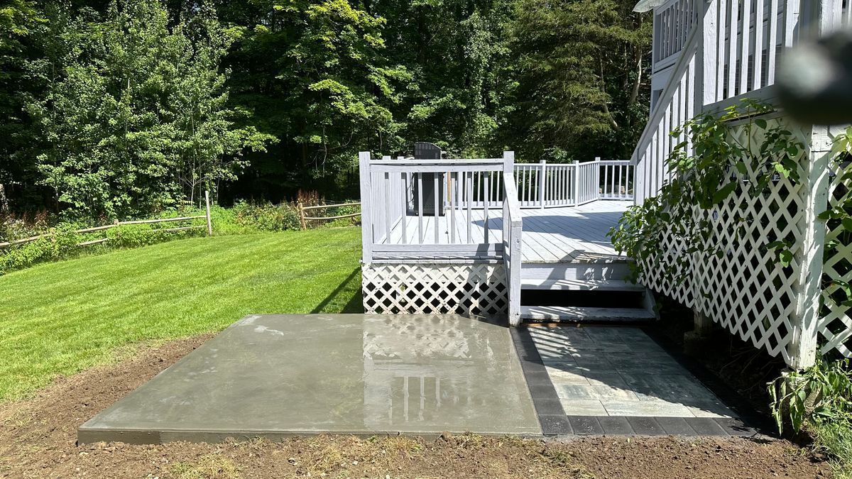 Backyard patio with a concrete pad and decorative brickwork, leading to a white deck. Green grass and trees surround the area.