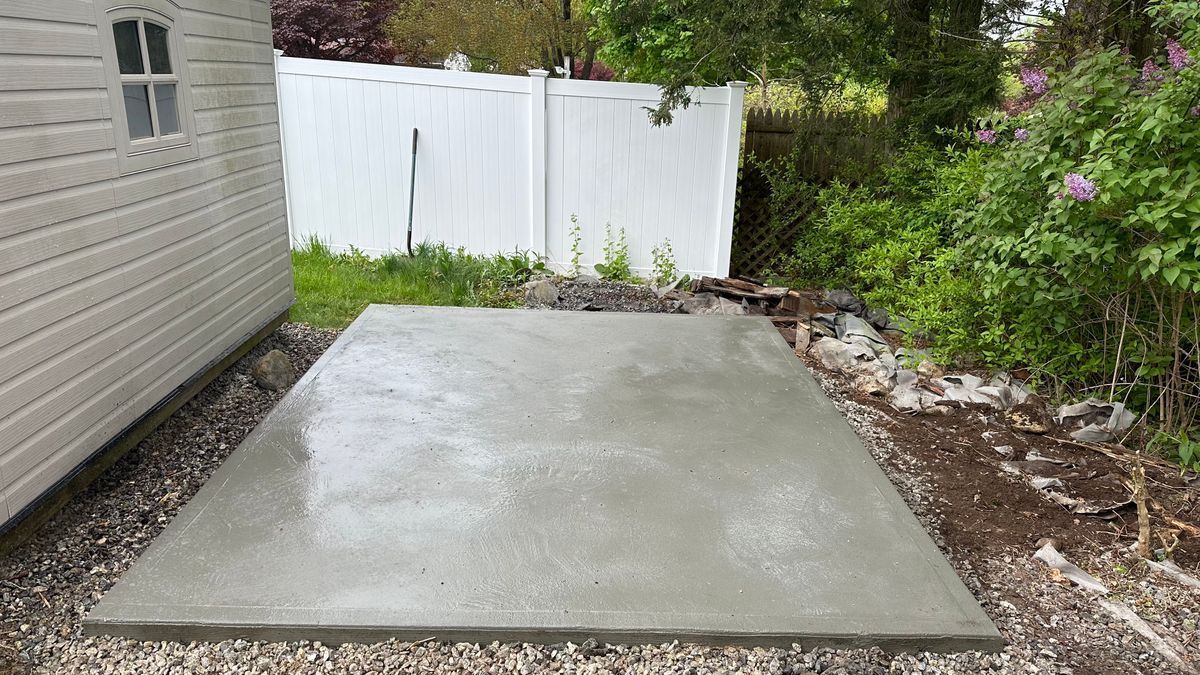 A freshly poured concrete pad next to a small shed and a white fence in a grassy backyard.