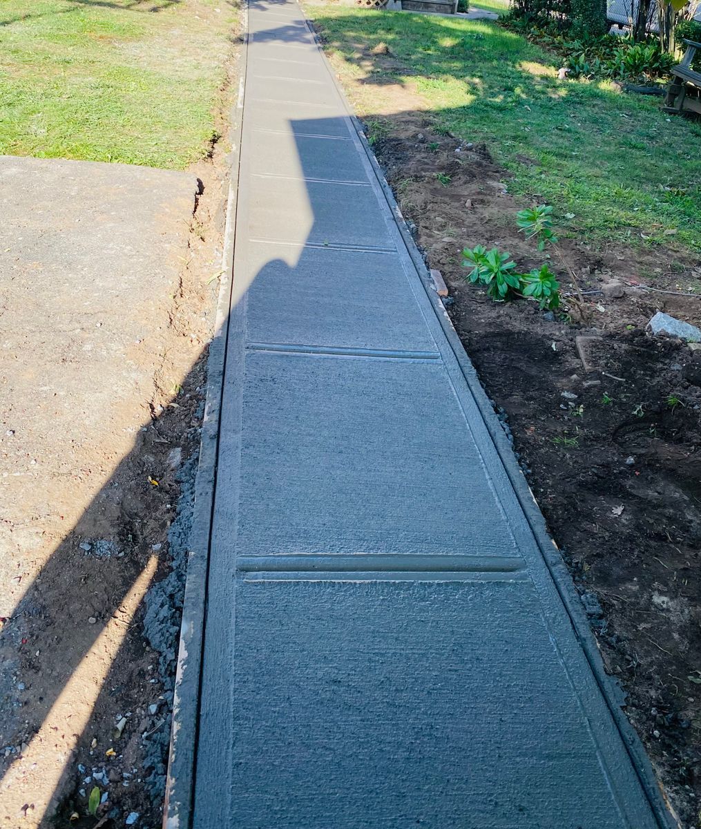 A newly poured concrete sidewalk with expansion joints, bordered by grass and soil.