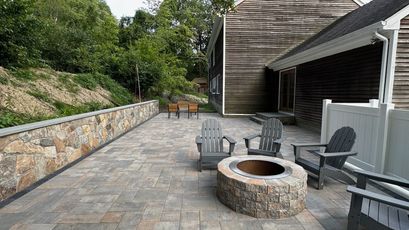 Stone pizza oven with a black door, built-in side counters, and chimney, set outdoors on a patio with a green lawn and trees.