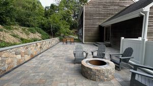 Stone pizza oven with a black door, built-in side counters, and chimney, set outdoors on a patio with a green lawn and trees.