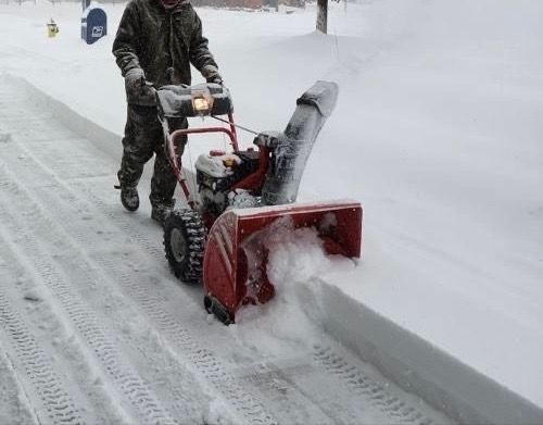 A person using a red snow blower on a snow-covered sidewalk.