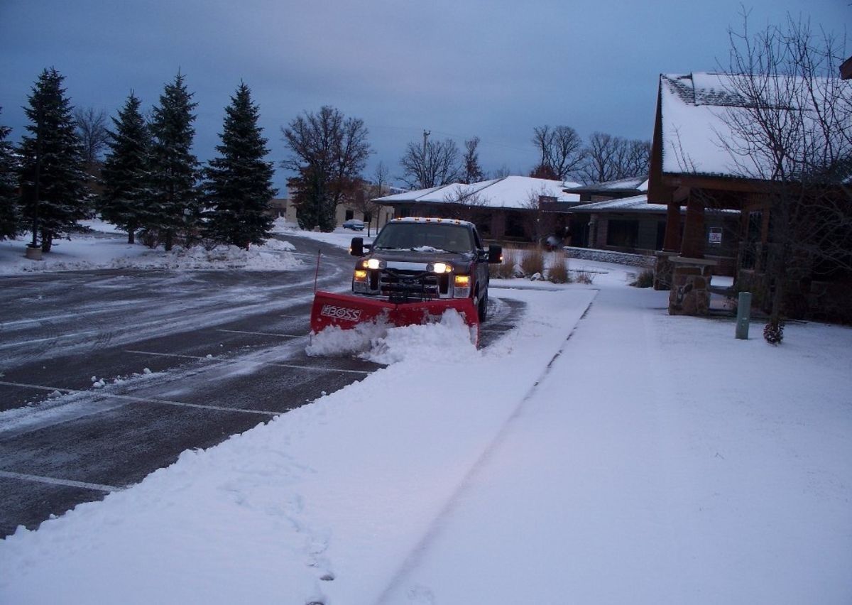 Snowplow clearing a snowy driveway in front of a building. Trees and sky in the background.