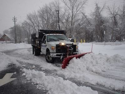 A white snowplow truck clearing a snowy road. The plow is red, and it pushes a large snowbank.