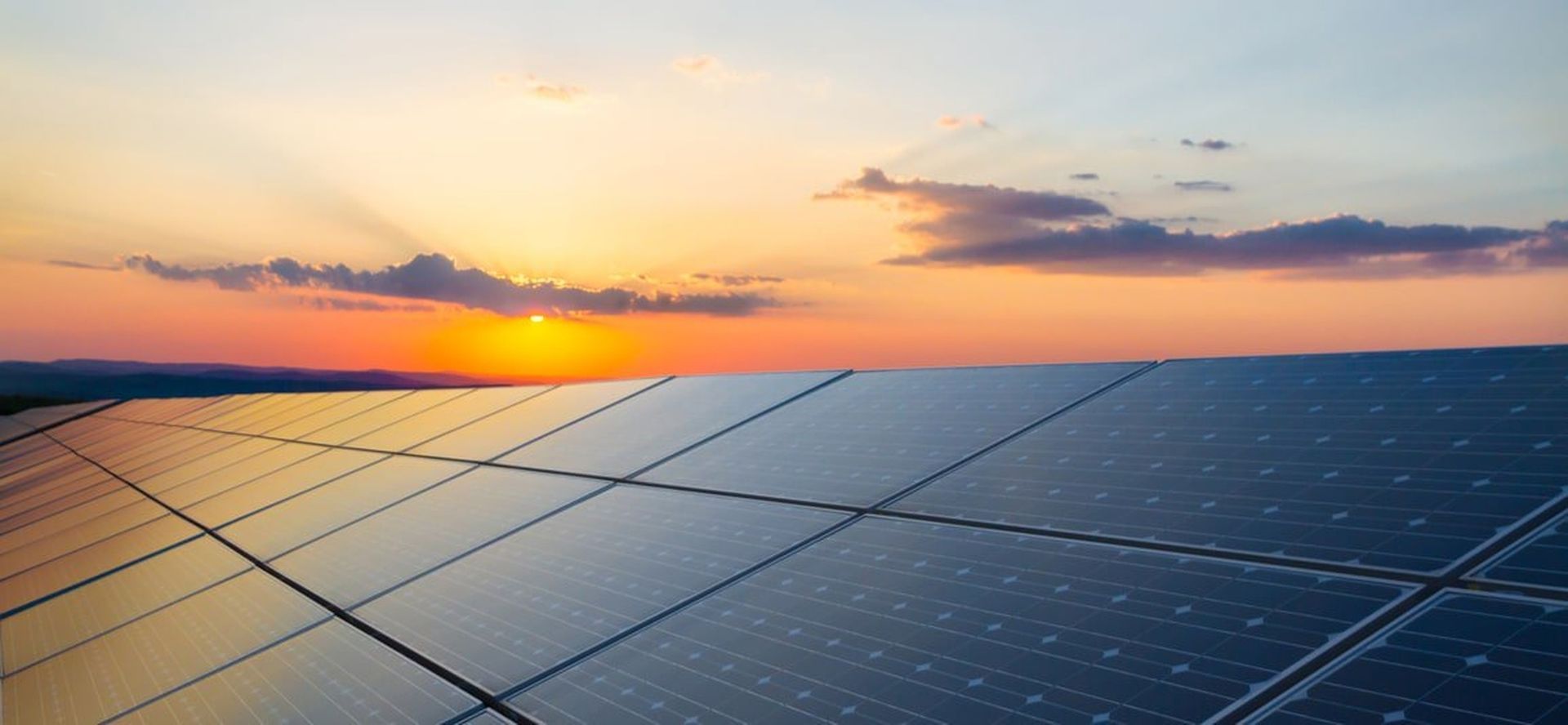 Solar panels at sunset under a colorful sky, with clouds and warm orange light.