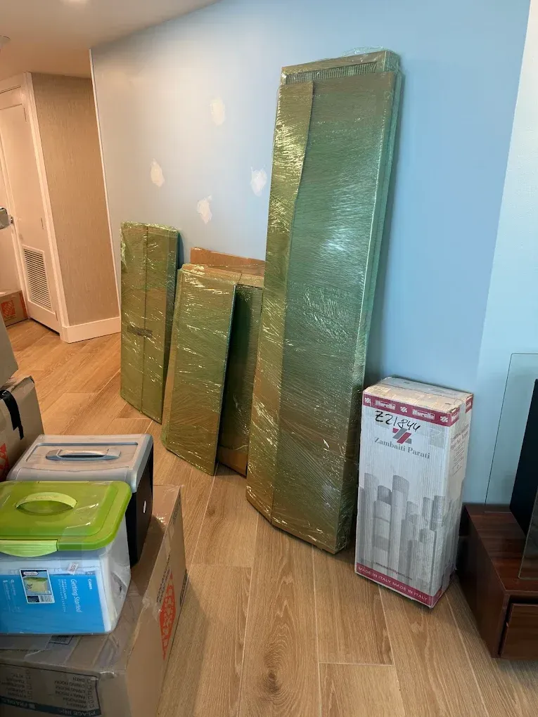 Wrapped furniture leaning against a light blue wall, boxes, and containers on a wooden floor.