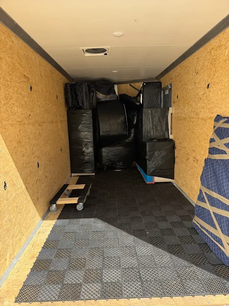 Interior of a cargo trailer loaded with wrapped furniture and a dolly, black flooring, and wood panel walls.