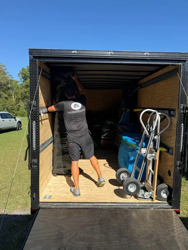 Man loading furniture into a black trailer, outdoors. A hand truck and blue items are also inside.