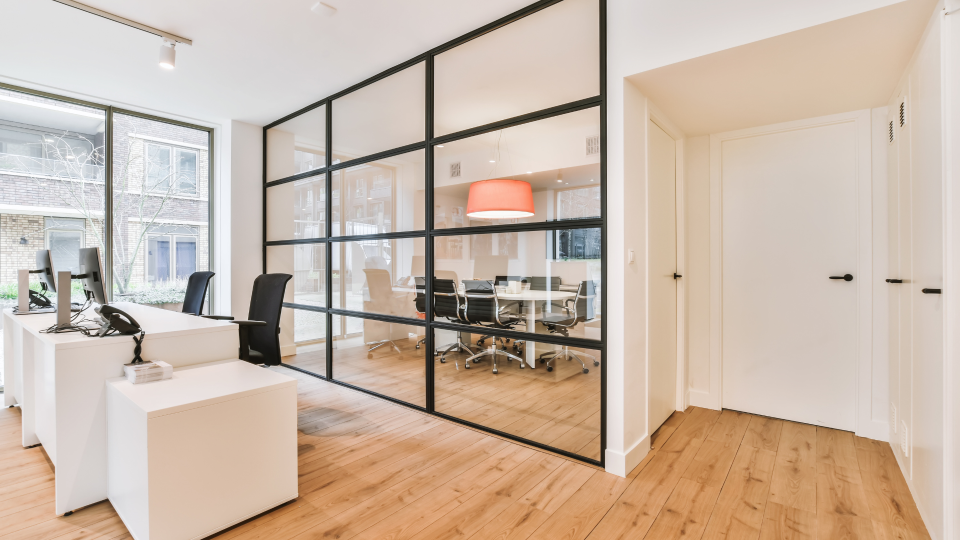 Office interior with glass-walled conference room, desks, wood floors, and a bright overhead light.