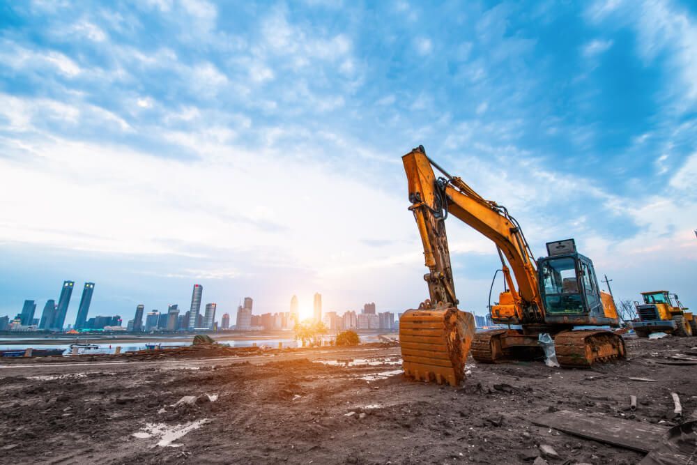 A Yellow Excavator Is Working On A Construction Site With A City Skyline In The Background — Dicko's Bobcat & Tipper in East Bendigo, VIC