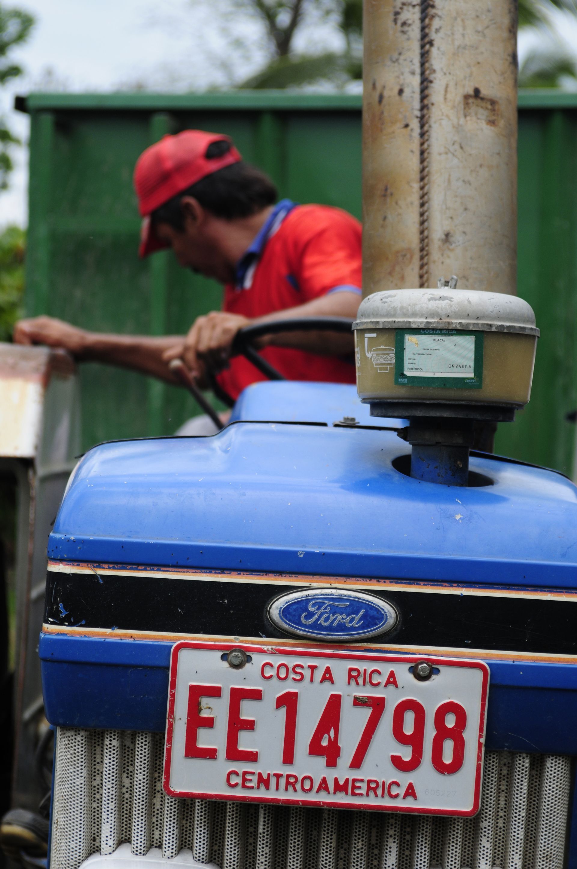 Man in rood shirt en pet rijdt op blauwe Ford-tractor in Costa Rica; kentekenplaat EE14798.