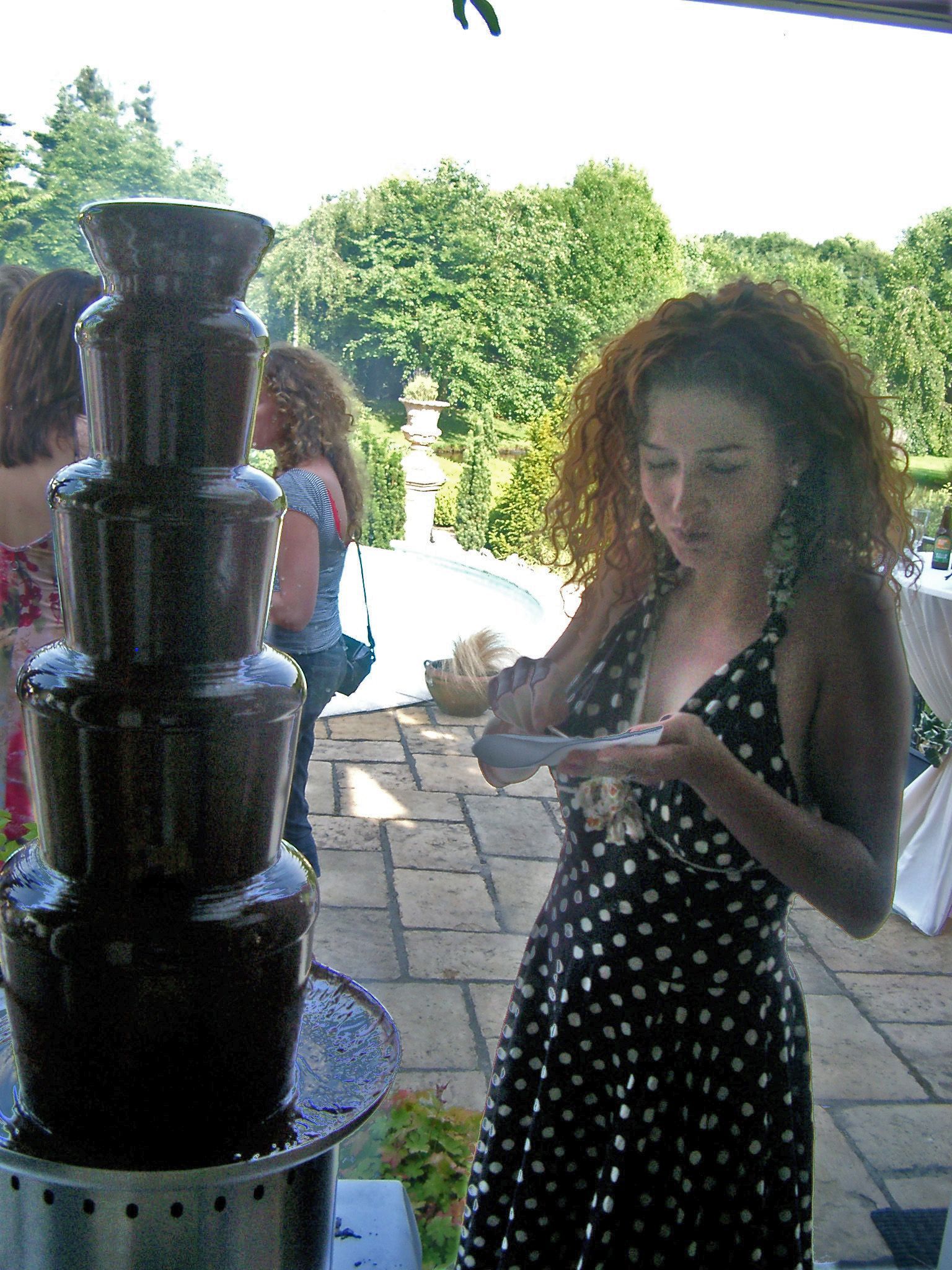 Vrouw in een jurk met stippen doopt eten in een chocoladefontein tijdens een buitenevenement. Groen gebladerte en een stenen terras op de achtergrond.