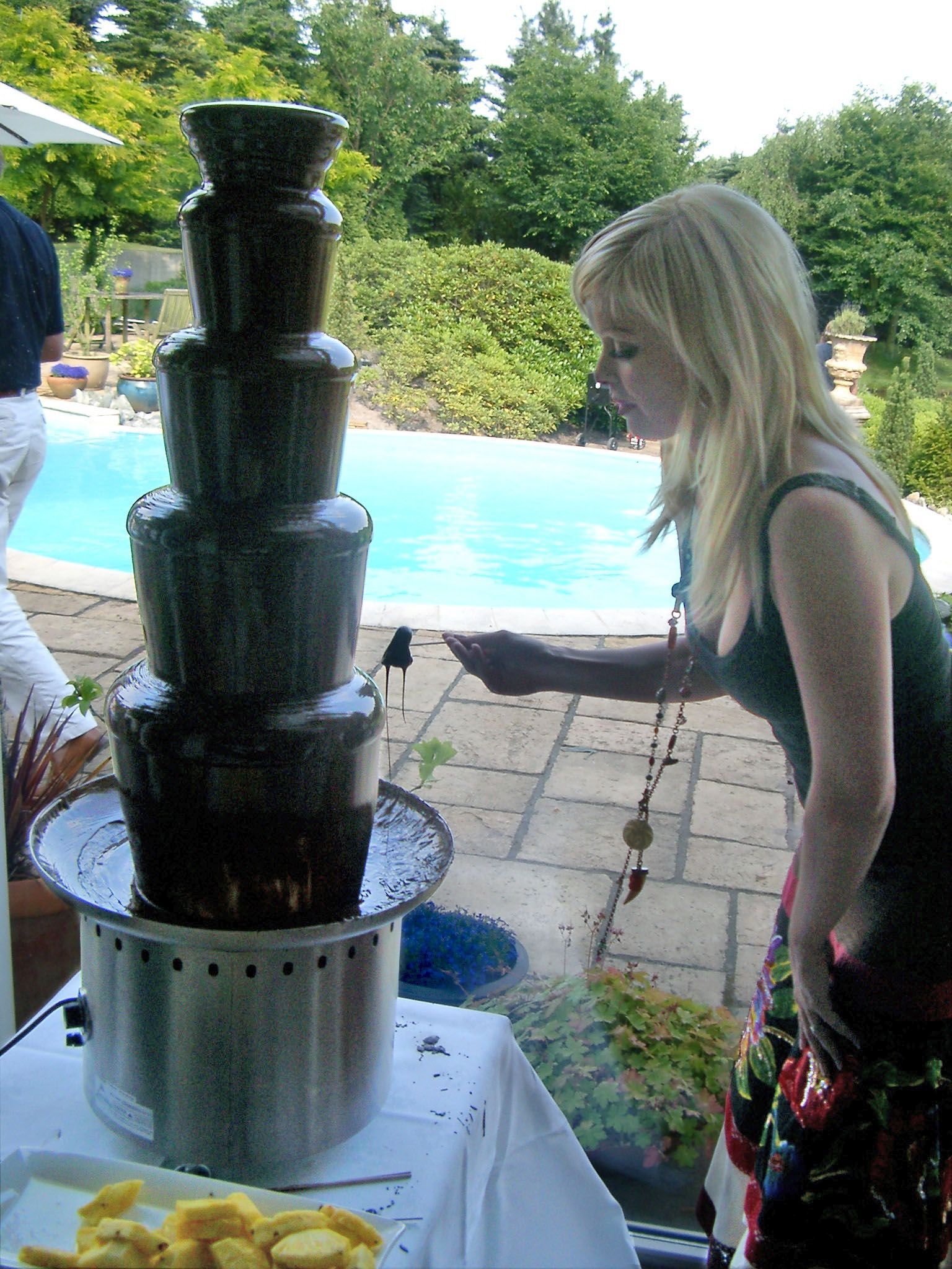 Een vrouw doopt een spiesje in een chocoladefontein bij een zwembad. Ze draagt een zwart tanktopje en een rok met print.