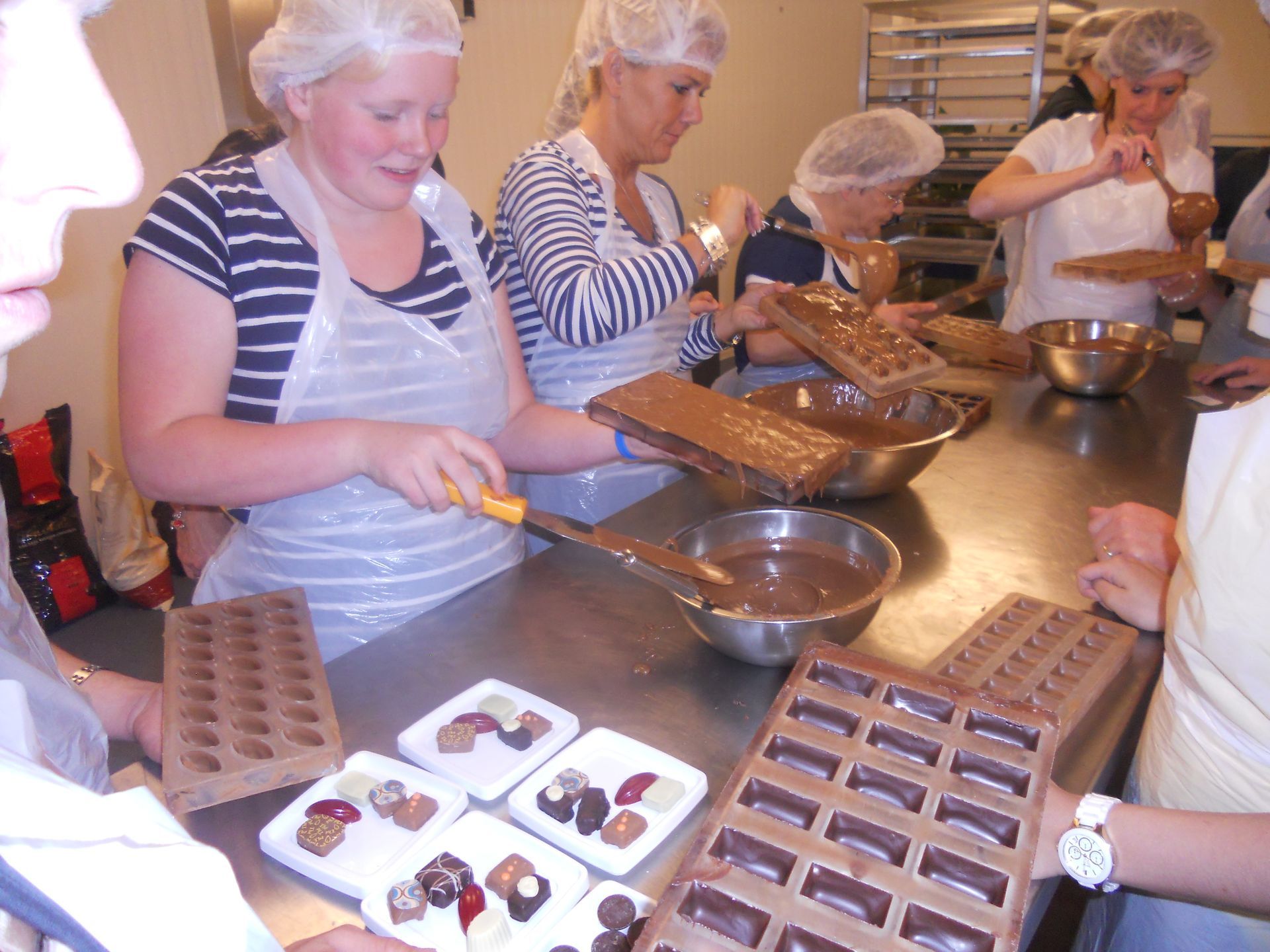 Mensen in haarnetjes en schorten maken chocolade in een keuken. Sommigen gieten gesmolten chocolade in mallen, terwijl anderen gevulde snoepjes versieren.