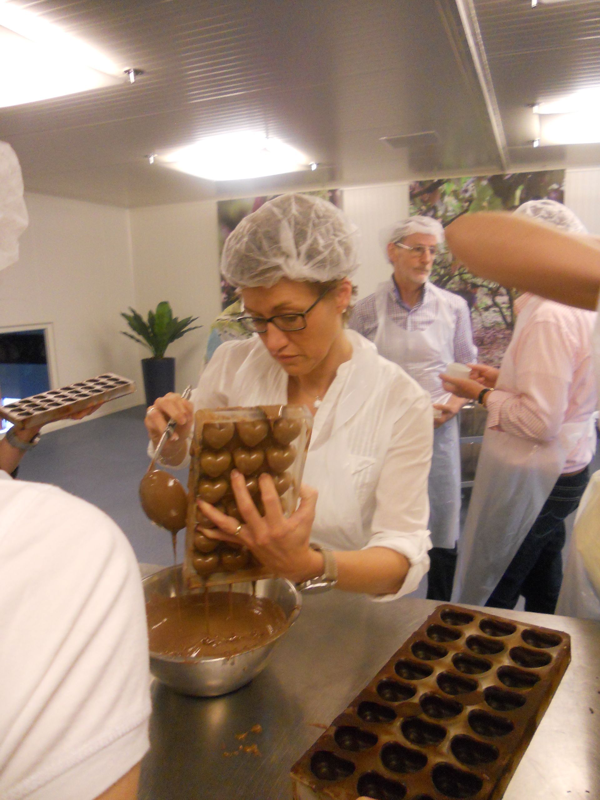 Een vrouw in haarnetje en labjas giet chocolade in een mal, waarschijnlijk in een professionele keuken. Andere mensen kijken toe.