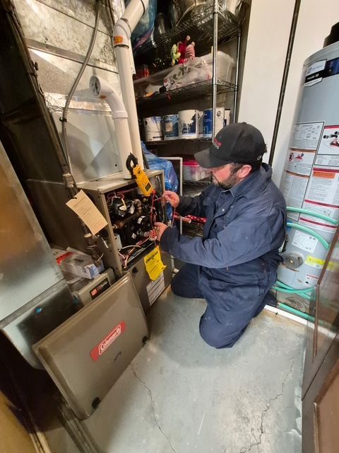 Technician kneels beside a furnace, adjusting controls in a utility room with pipes and storage shelves.