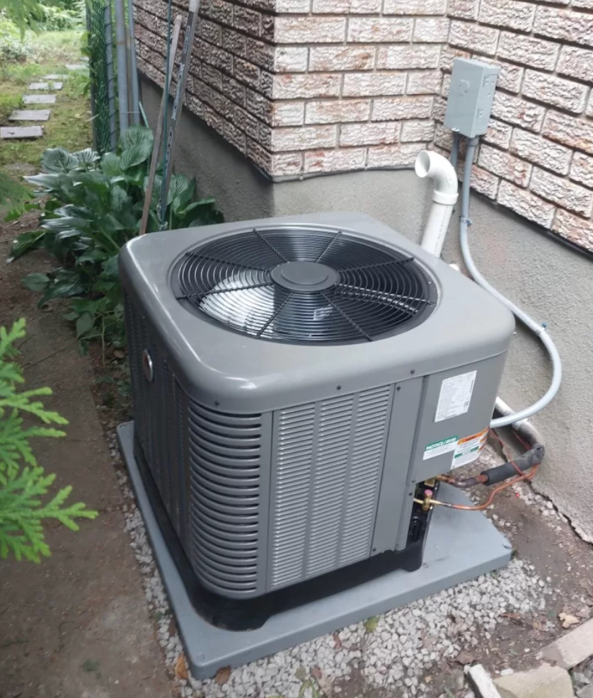 Outdoor air conditioning unit beside a brick house, with a fan on top and white conduit pipe.