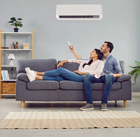 Couple relaxing on a gray sofa, using a remote in a bright living room with a wall-mounted air conditioner.