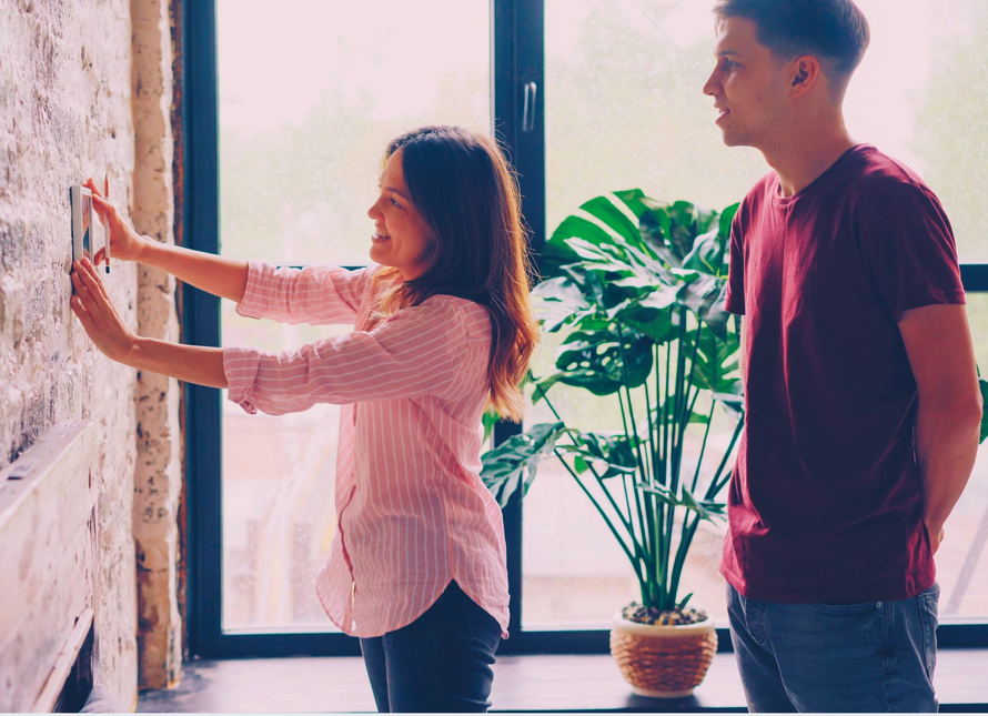 Two people near a window, arranging paper on a wall beside a potted plant.